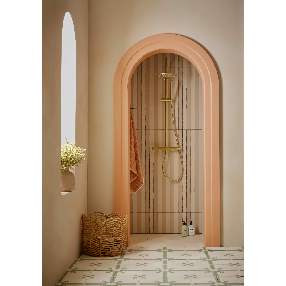 Modern bathroom with arched terracotta doorway featuring brushed brass shower system, vertical tile surround, woven basket storage, potted plant in wall niche, and patterned floor tiles in neutral tones