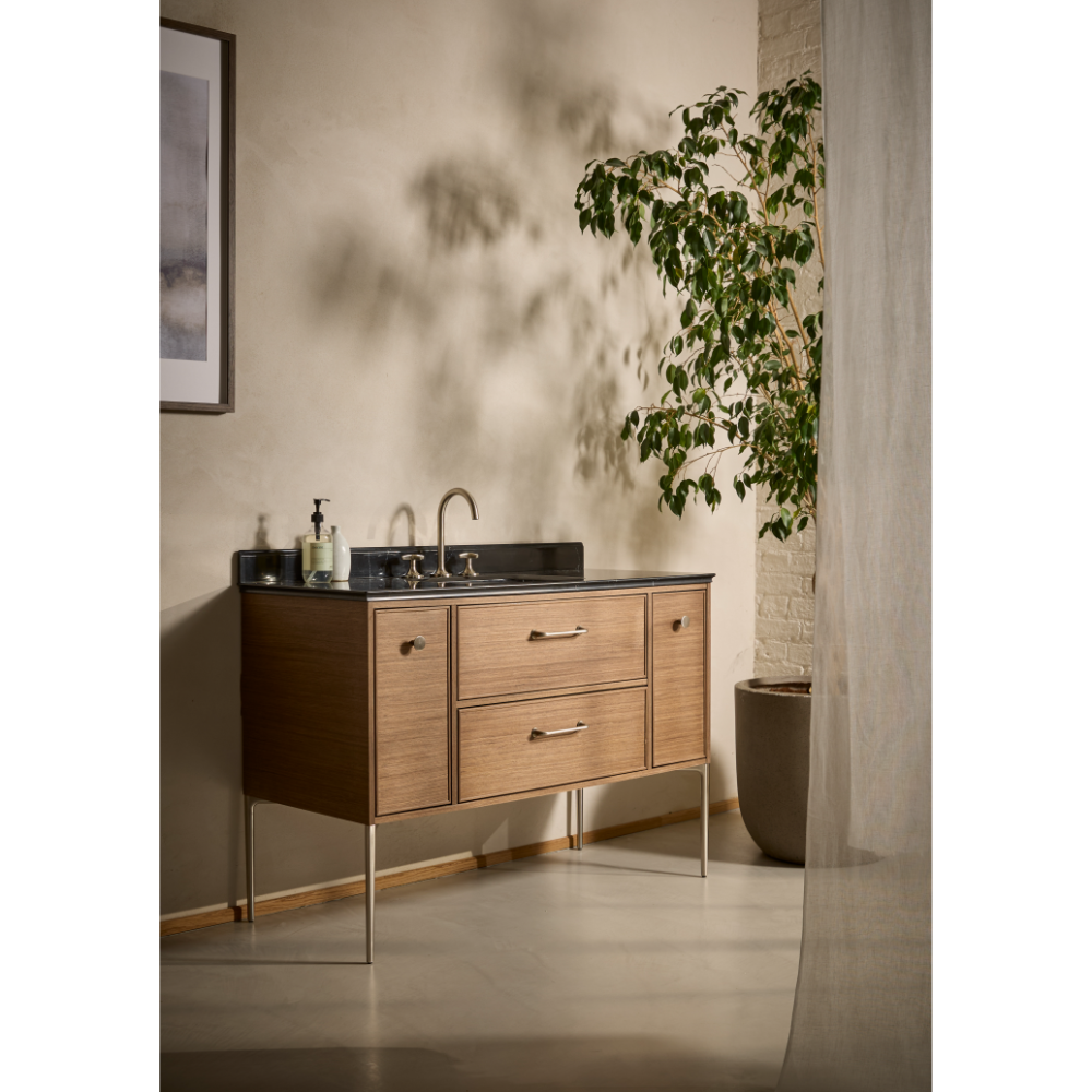 Light oak bathroom vanity unit with dark countertop and bronze fixtures, featuring two drawers and cabinet storage, set against white wall with climbing ivy plant and concrete column detail
