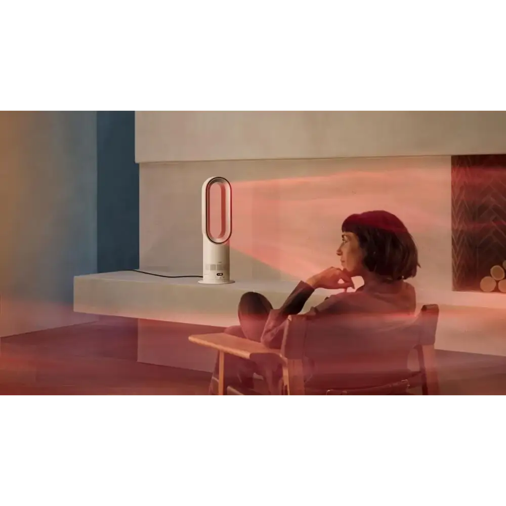 Woman sitting in pink armchair in modern minimalist room with warm ambient lighting, operating white and silver Dyson Hot + Cool HF1 heater fan on table