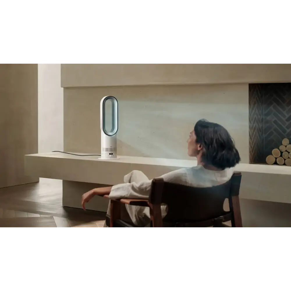 Woman sitting at desk facing white and silver Dyson Hot + Cool HF1 heater fan in minimalist beige interior with wooden chair and shelving