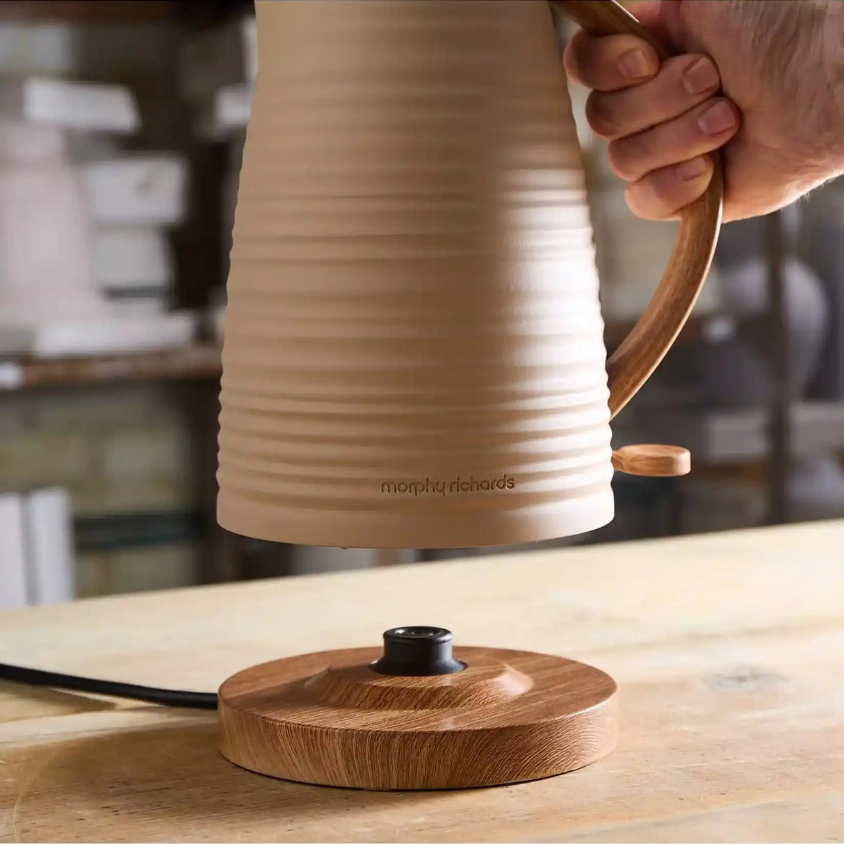 Morphy Richards Clay Felspar electric jug kettle in cream and wood finish being held over its matching wooden base, displaying the modern design with brown wood-effect handle and accents on a kitchen countertop