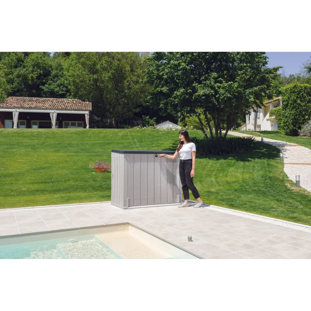 Woman opening a taupe grey and anthracite Toomax Storer Plus garden storage unit on a patio beside a swimming pool, with manicured lawn and residential buildings in the background