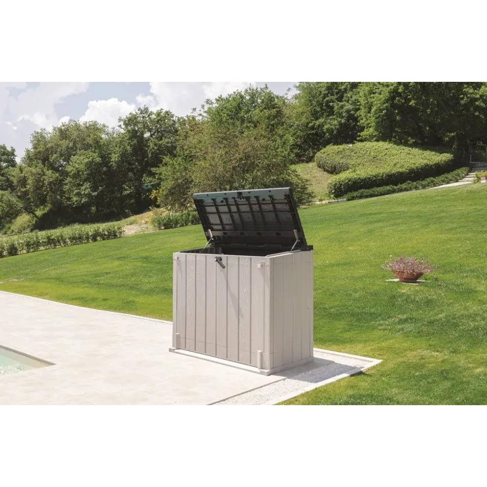 Toomax Storer Plus taupe grey and anthracite garden storage unit with open lid displayed on a patio beside a manicured lawn with hedges and trees in the background