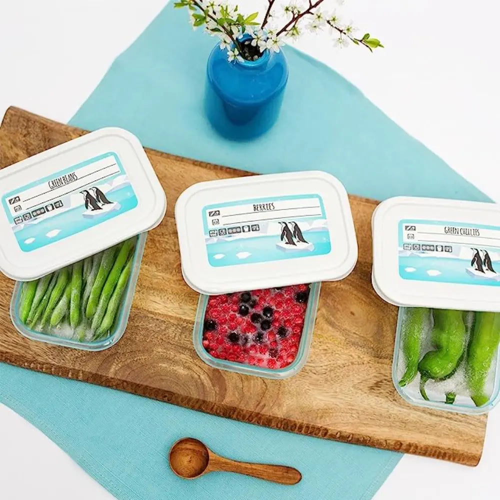 Three Keeeper food storage containers with turquoise lids arranged on a wooden cutting board atop a light blue surface, containing green beans, mixed berries, and snap peas, with a blue vase of white flowers and wooden spoon visible, displaying product labeling on white lids