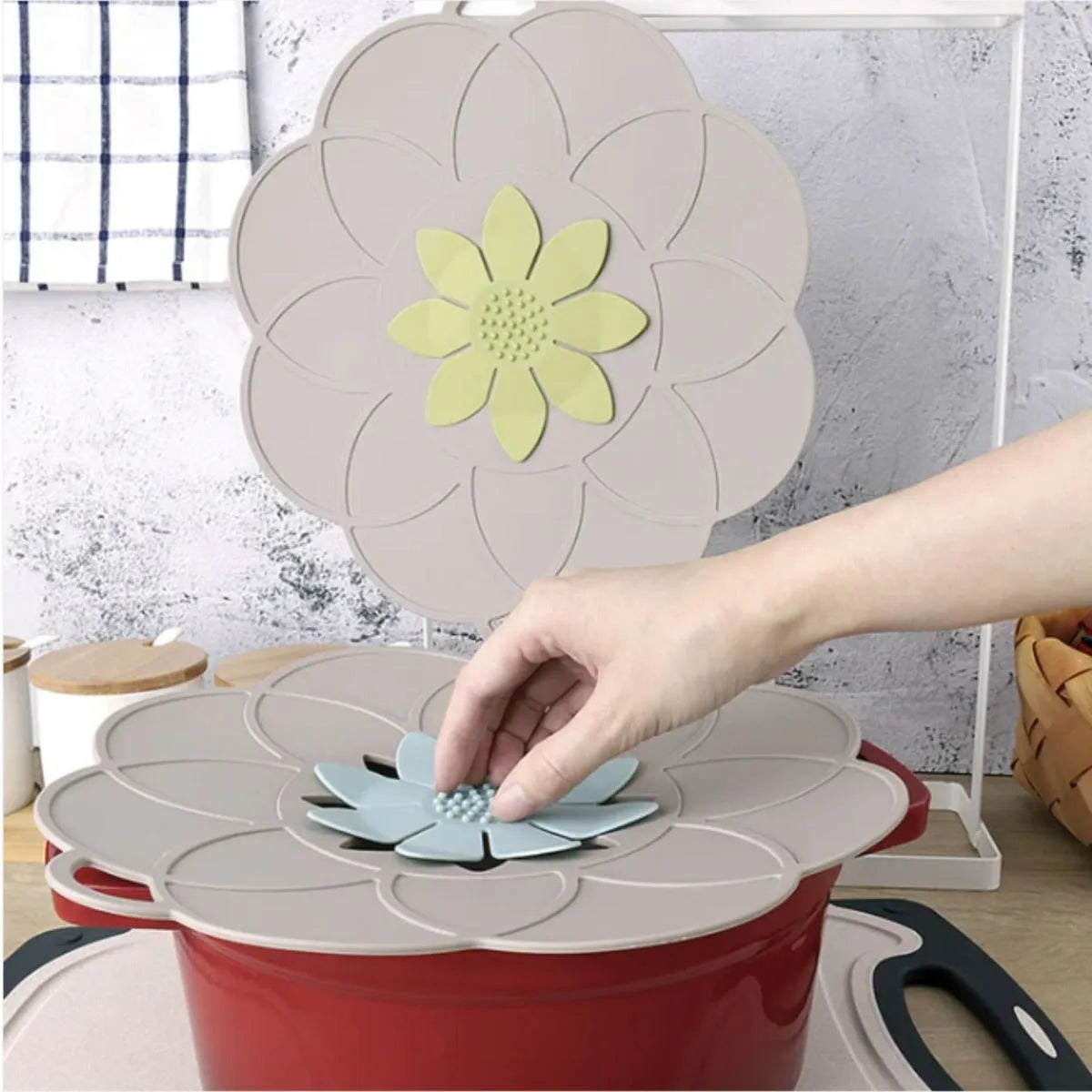 Flower-shaped silicone lids in various colors including red, cream, and blue displayed on cookware, with a hand placing a light blue petal-designed lid on a red pot, showing the anti-spill functionality of the Kitchen Academics silicone lids