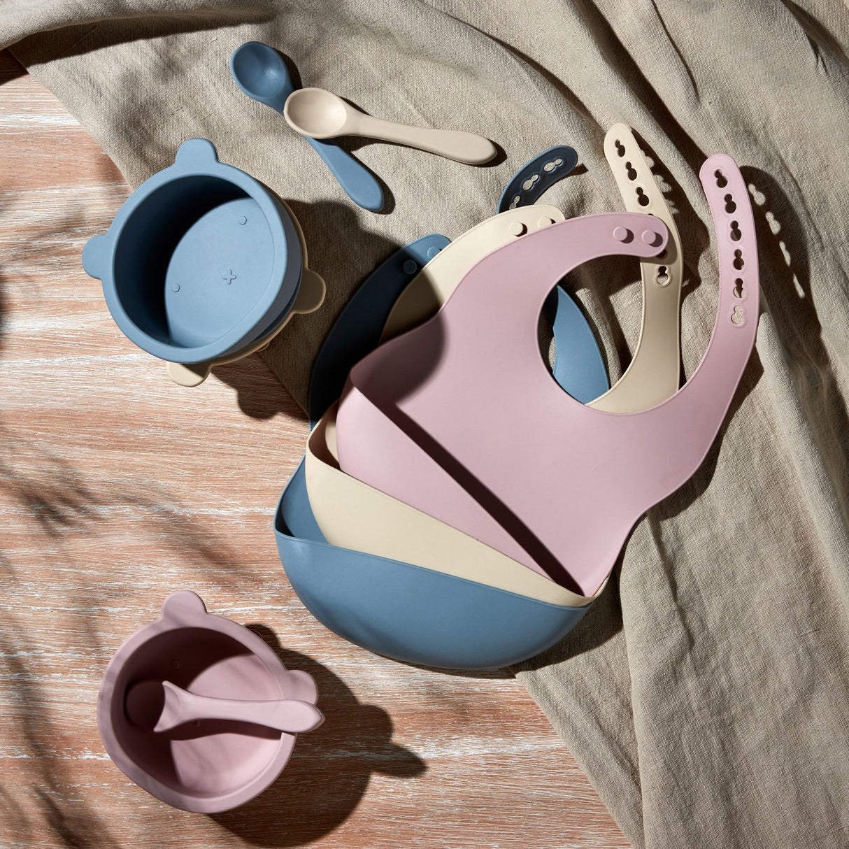 Bambino silicone feeding set in pink, blue and cream displayed on wooden surface with beige fabric, including bibs with snap buttons, bowls with handles, and matching spoons