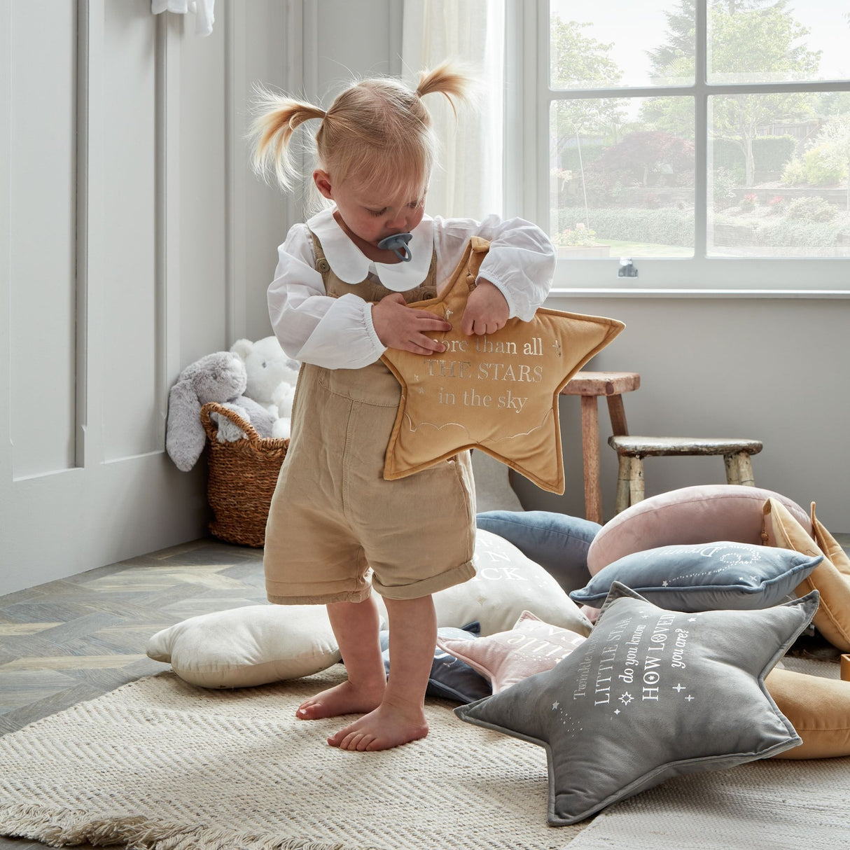 Toddler playing with gold velvet star-shaped cushion featuring "Twinkle Little Star" text in a bright nursery room with additional pastel star pillows on floor, wicker basket with stuffed animals, and large windows overlooking garden