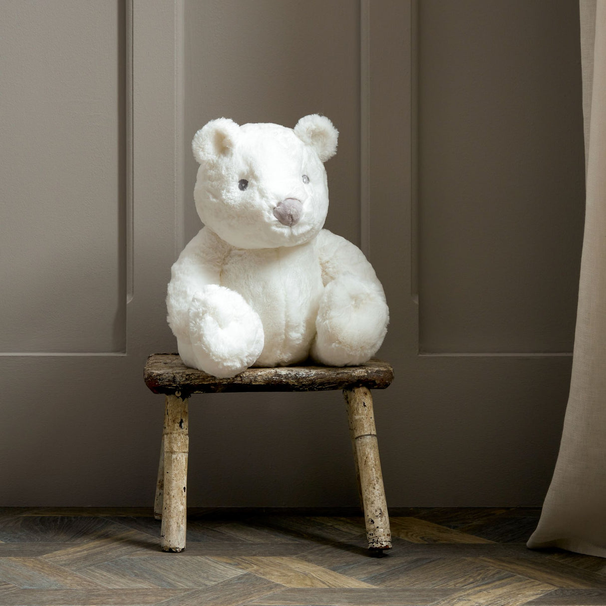 Large white plush teddy bear sitting on rustic wooden stool against neutral beige wall backdrop