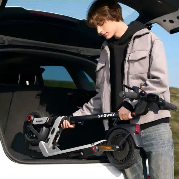 Young person holding a black and white Segway Ninebot E3 Pro electric scooter by an open car trunk on a sunny day with clear blue sky