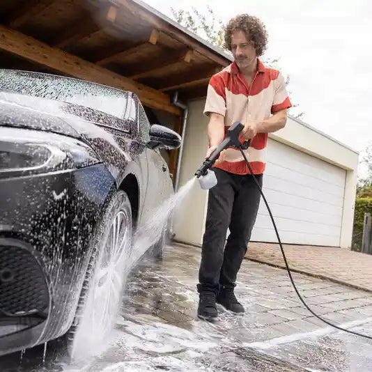Man in red and cream shirt using Karcher K3 pressure washer with blue hose to clean black car in driveway, with garage in background