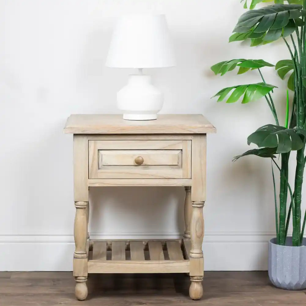 Light wood one drawer end table with turned legs and slatted shelf, topped with white ceramic table lamp, styled with potted green plant against white wall