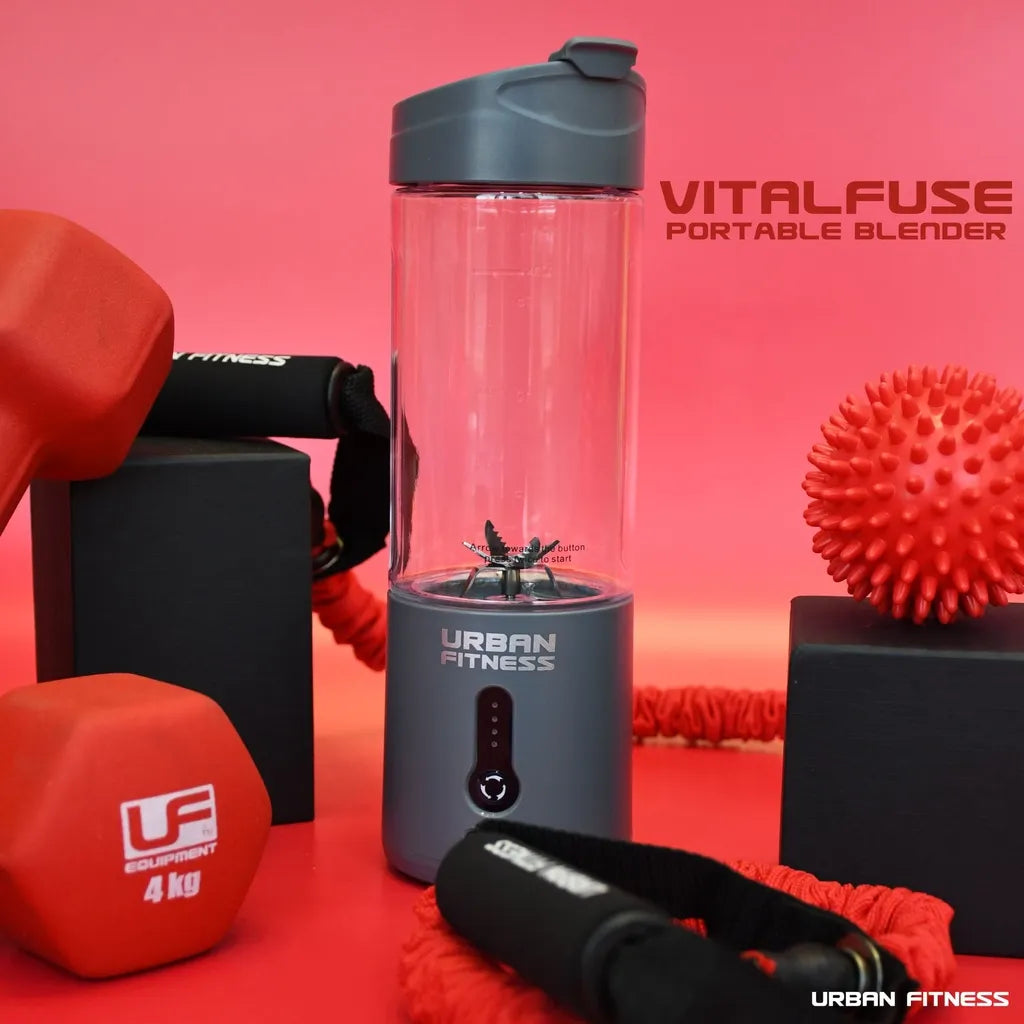 Urban Fitness VitalFuse portable blender in grey with black lid surrounded by red fitness equipment including dumbbells, resistance ball and jump rope on pink background