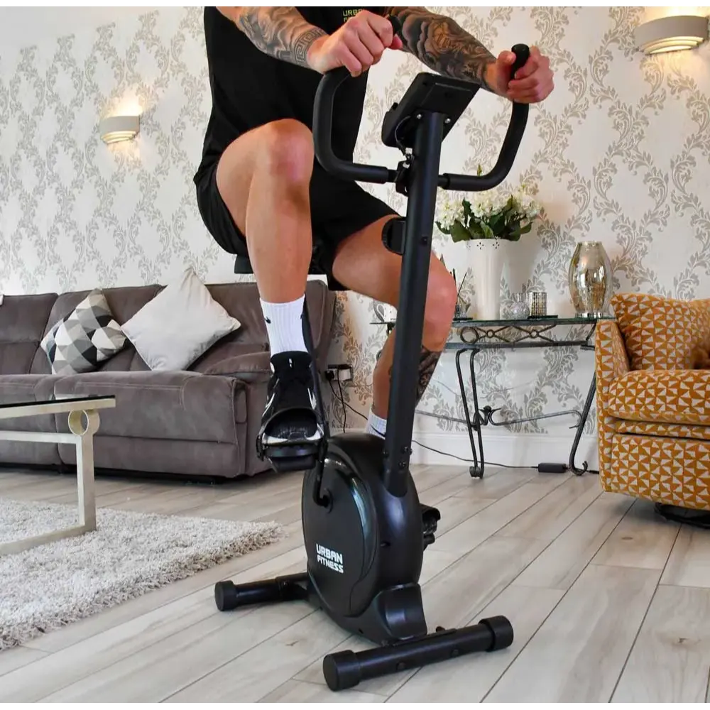 Person using Urban Fitness black magnetic exercise bike in modern living room with grey sofa, patterned wallpaper, and wooden flooring