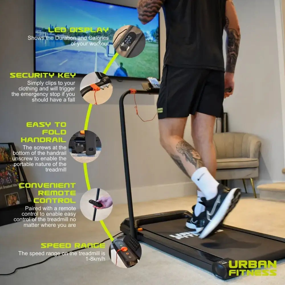 Man running on Urban Fitness black and white walking treadmill in living room with TV screen displaying workout stats, showing product features including LED display, security key clip, foldable handrail, remote control, and speed range of 1-8km/h with yellow text annotations