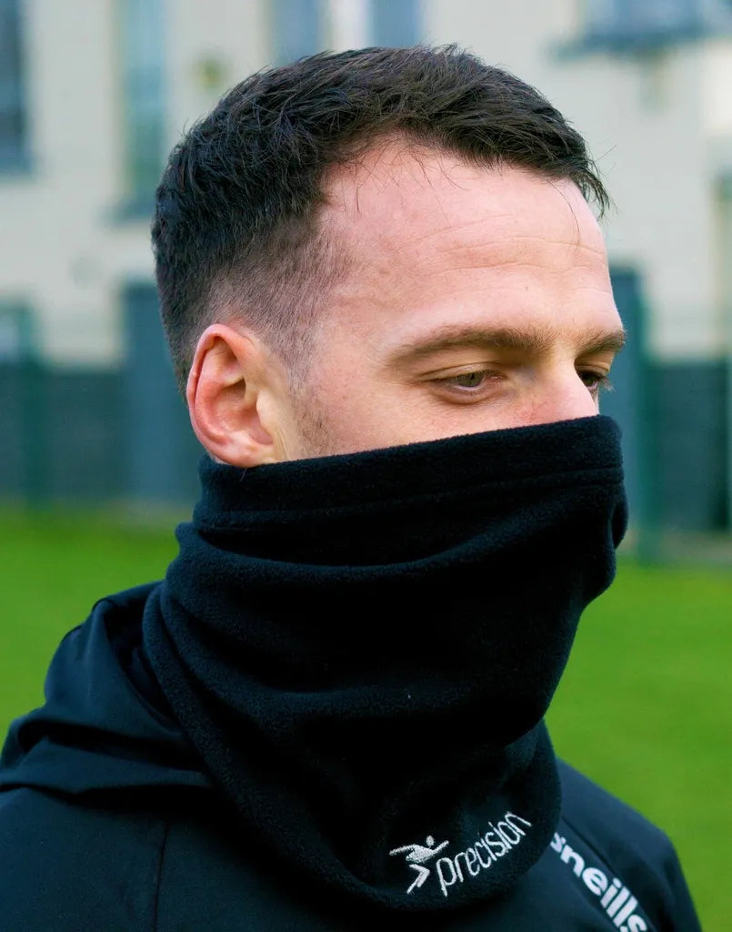 Man wearing black Precision Essential Neck Warmer Snood with logo text, styled as high neck gaiter, outdoor setting with grass and building background