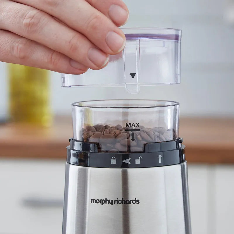 Hand holding transparent water reservoir above Morphy Richards stainless steel electric coffee grinder with black control band and coffee beans visible in grinding chamber marked with MAX line