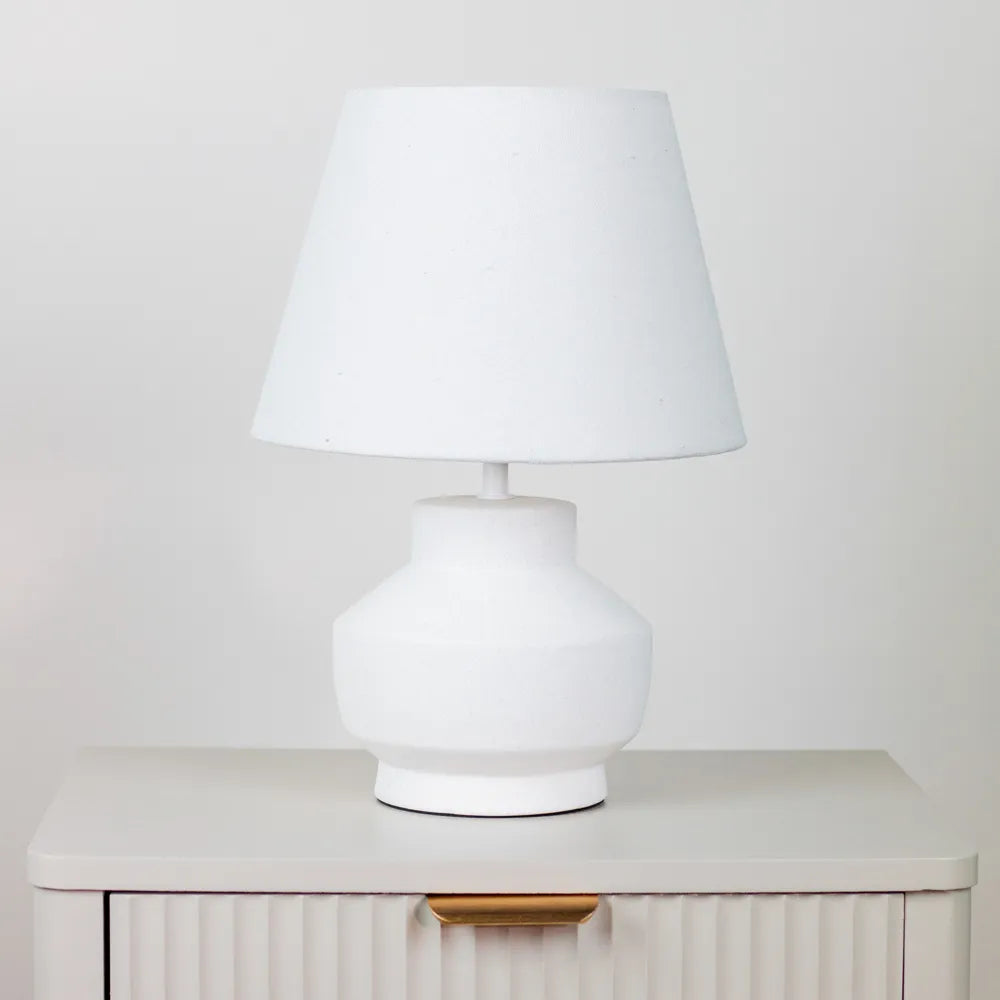White ceramic table lamp with bulbous base and conical linen shade sitting on cream ribbed bedside cabinet against neutral background