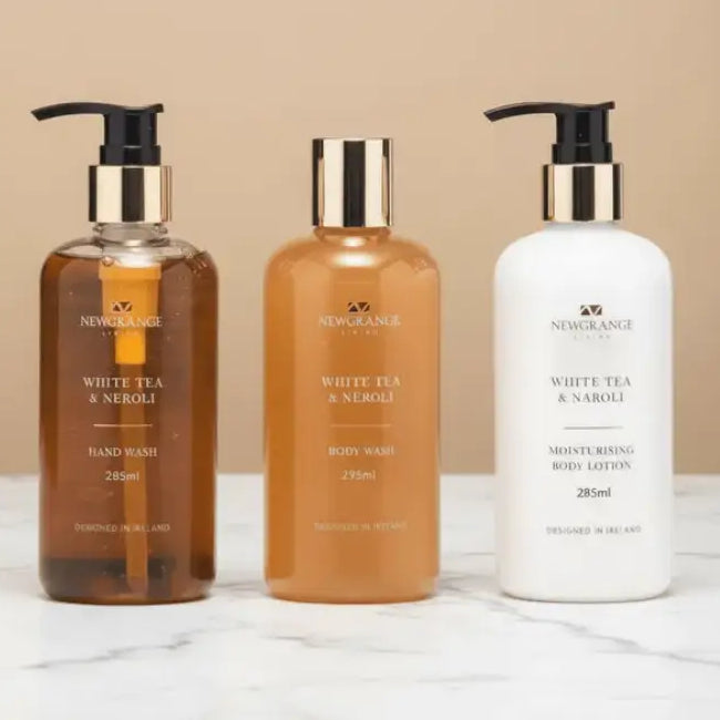 Three-piece Newgrange Living White Tea and Neroli body care set displayed on marble surface featuring amber glass hand wash bottle, orange glass body wash bottle, and white lotion bottle, each 285ml with black pump dispensers and gold-toned caps, designed in Ireland