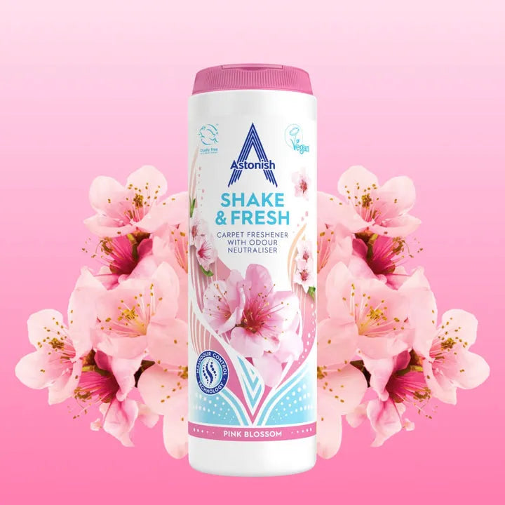 Astonish Shake and Fresh Pink Blossom carpet freshener 400g bottle with white container and pink lid, surrounded by pink and magenta flowers on pink background, showing product packaging with vegan certification logo
