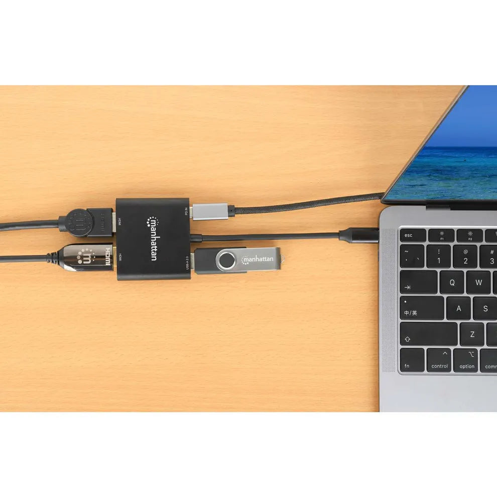 Manhattan USB-C to Dual HDMI multiport adapter with 100W power delivery connected to MacBook Pro on wooden desk, showing black adapter hub with multiple cables and USB flash drive