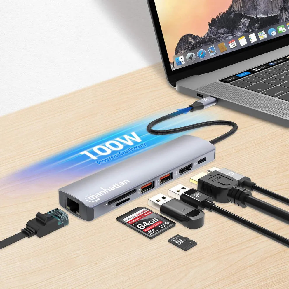 Manhattan USB-C 7-in-1 docking station with power delivery in silver aluminum connected to MacBook Pro via blue cable, displaying multiple ports including HDMI, USB-A, SD card reader, and Ethernet, alongside USB cables and memory cards on wooden desk surface