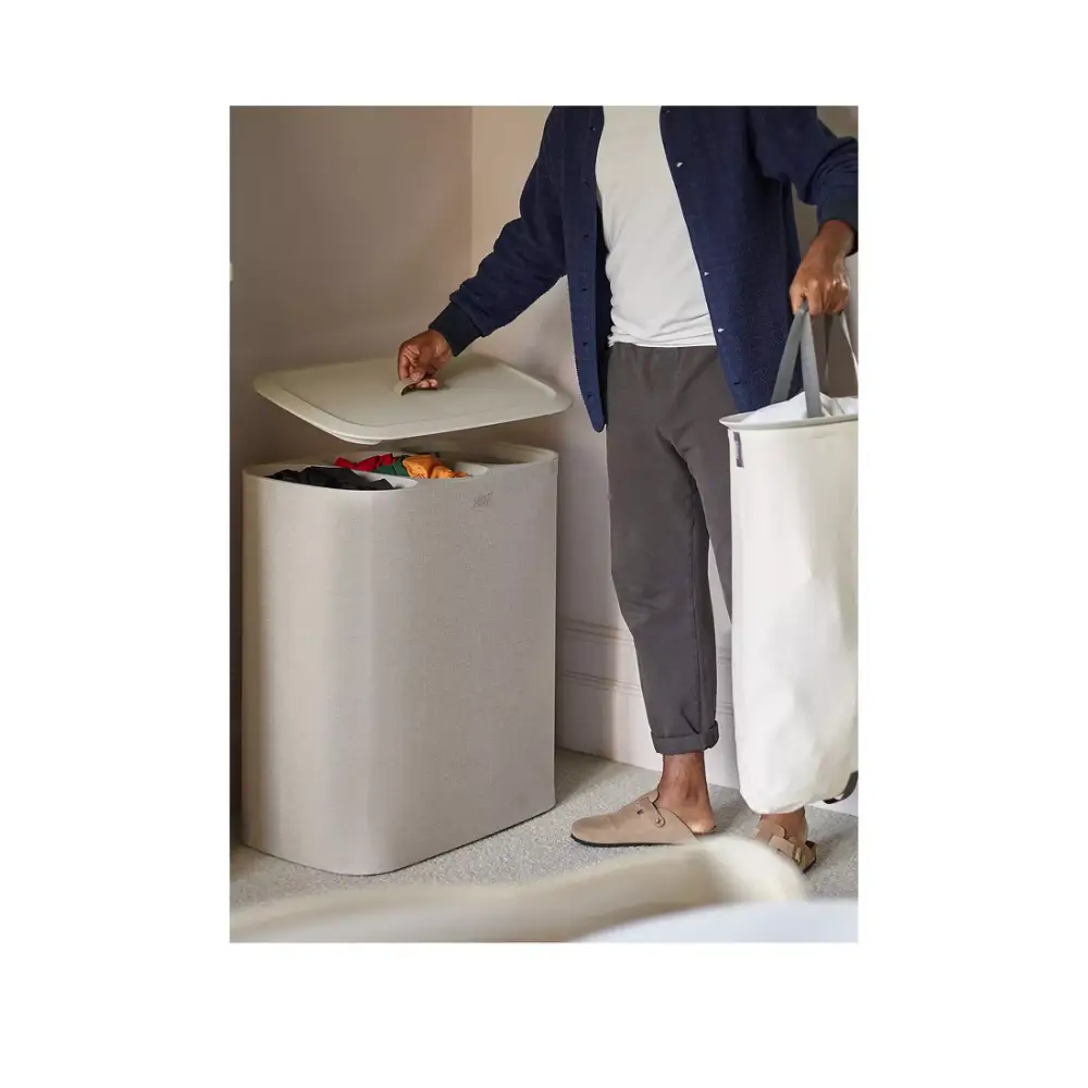 Person in navy cardigan and grey trousers placing colored clothing into a white cylindrical laundry basket with lid, alongside matching white laundry baskets on a light floor