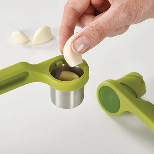 Hand using green and stainless steel Helix garlic press to crush garlic clove over white surface with garlic pieces visible in background