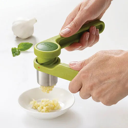 Green and stainless steel Helix garlic press being used to crush garlic clove over white bowl, with fresh garlic and mint leaves visible on white background