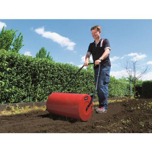Man using a red metal garden roller on soil in a sunny garden with green hedges and blue sky in the background