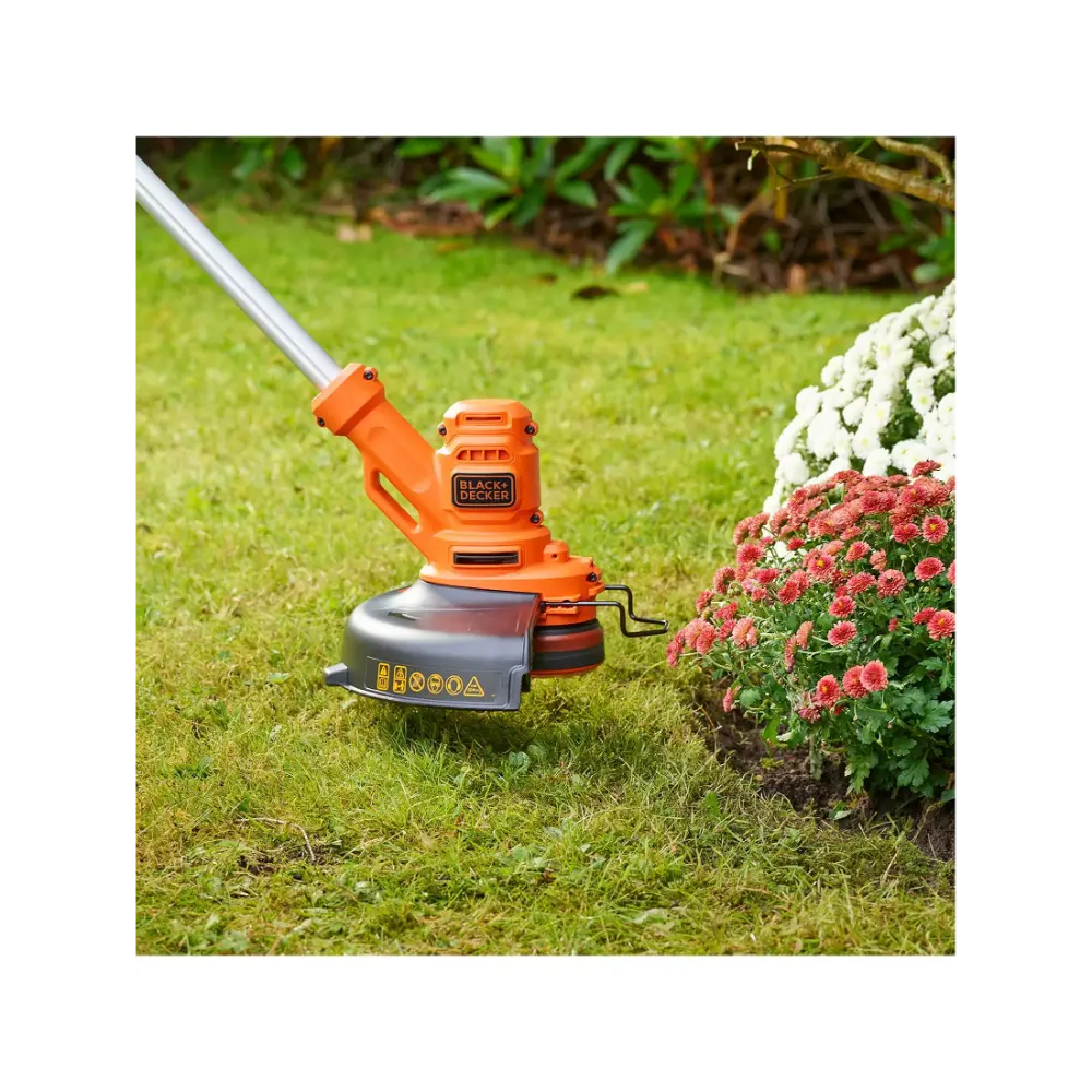 Black & Decker BESTA525 orange and grey electric grass strimmer with cutting head positioned on lawn next to pink and white flowering plants in garden setting