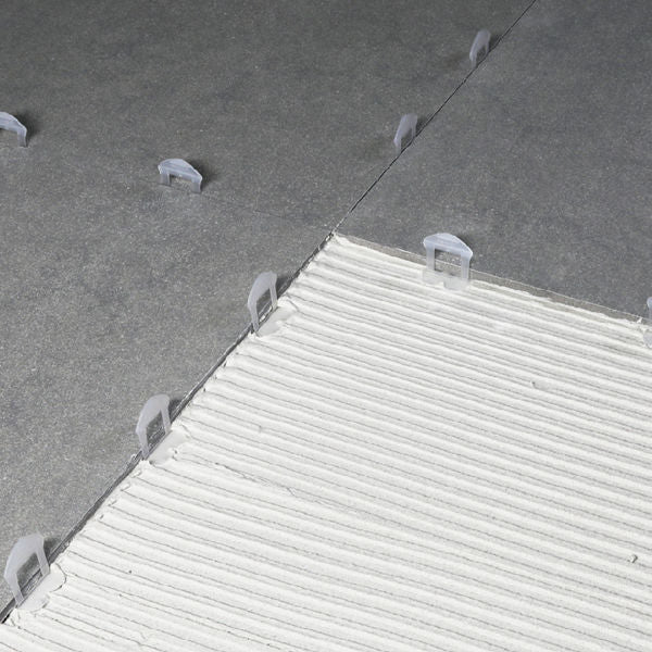 Overhead view of white plastic tile levelling wedge clips installed on gray floor tiles during installation, showing spacer positioning and alignment system