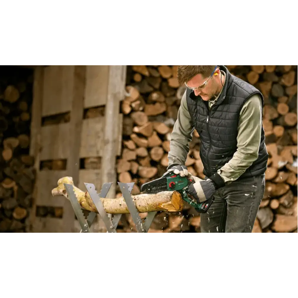Man using Bosch EasyChain 18V cordless chainsaw to cut log on sawhorse with stacked firewood in background
