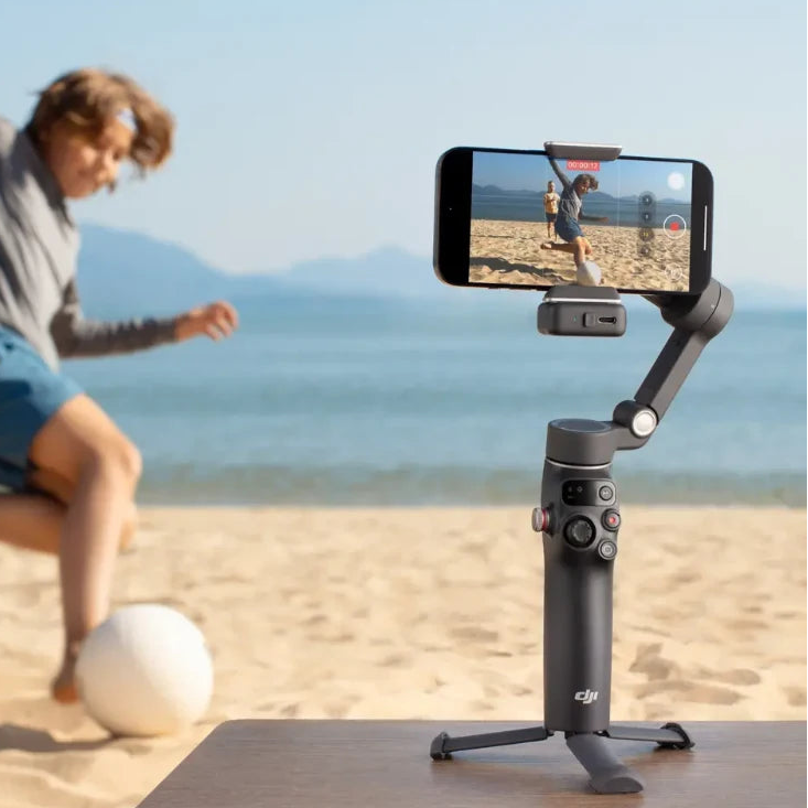 DJI Osmo Mobile 7P Pro smartphone gimbal in dark grey mounted on wooden table recording beach scene with children playing soccer, demonstrating video stabilization technology for mobile content creation