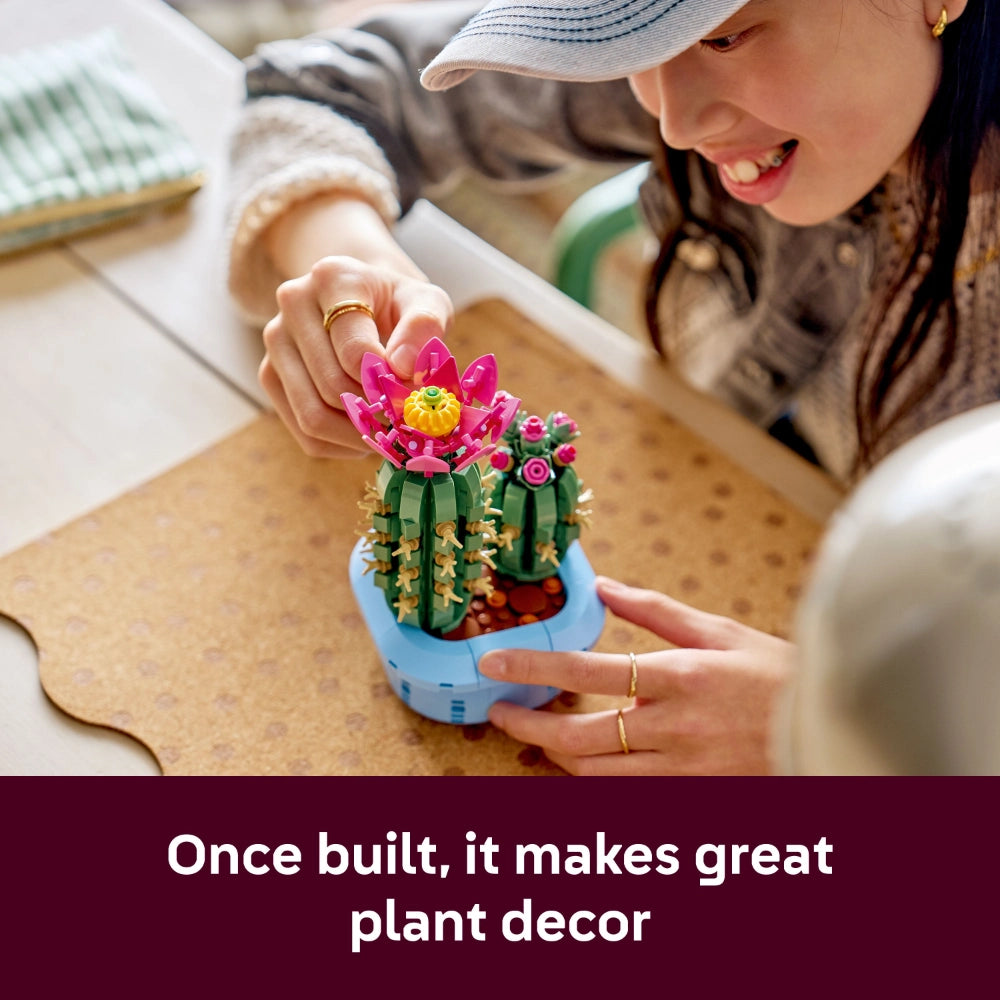 A young woman wearing a blue and white striped cap smiles while touching a LEGO Flowering Cactus set displayed in a light blue pot on a cork surface, featuring bright pink and yellow blooms with green segmented cactus stems, positioned as decorative home plant decor