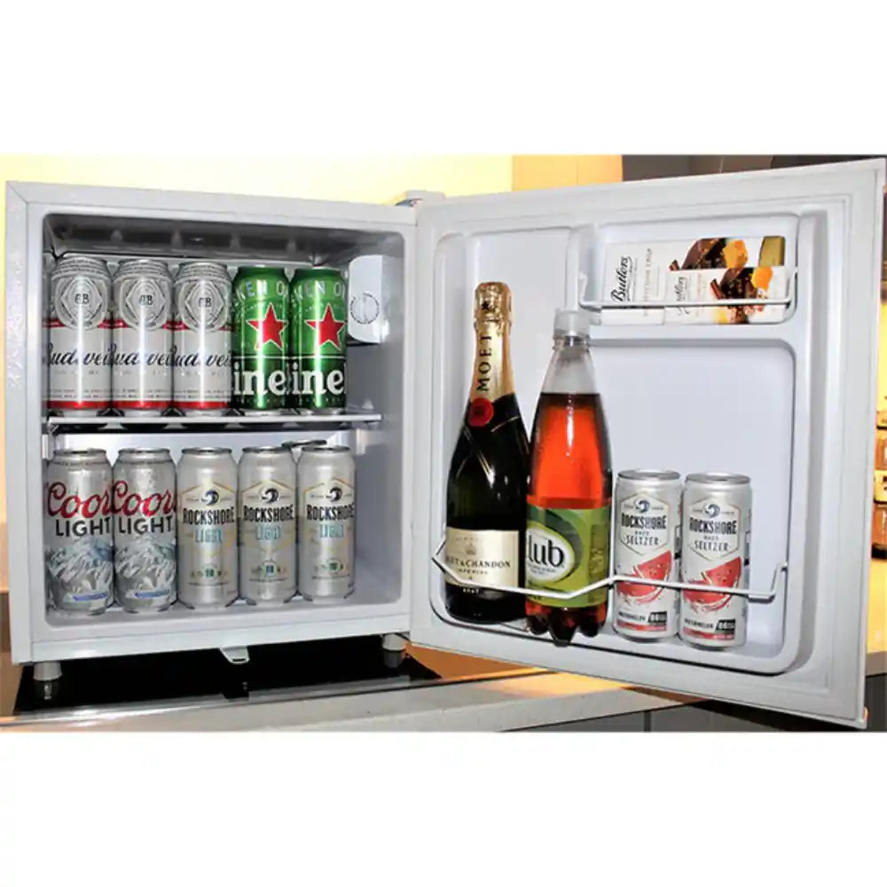 White compact tabletop refrigerator with open doors displaying beverages including beer cans, champagne bottles, and soft drinks on white shelving against a yellow and brown wall background