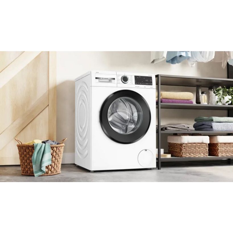 White Bosch Series 6 washing machine with black drum door in modern laundry room setting, surrounded by wicker laundry basket, wooden cabinetry, and storage shelves with folded towels and plants