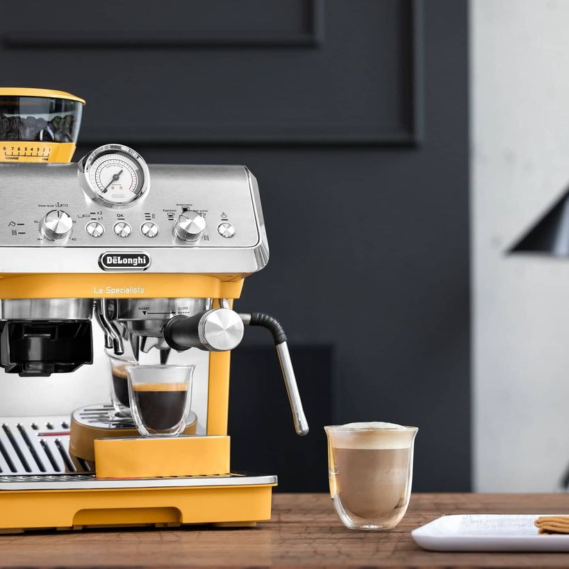 DeLonghi La Specialista Arte manual bean to cup espresso machine in yellow and stainless steel with dual espresso shots being dispensed into glass cups on wooden counter with dark background