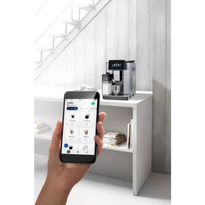 Hand holding smartphone displaying DeLonghi coffee machine app with various espresso and cappuccino options, positioned in front of the DeLonghi PrimaDonna Soul automatic coffee machine in metal black finish sitting on a white shelf in a minimalist white interior with stairs visible in background