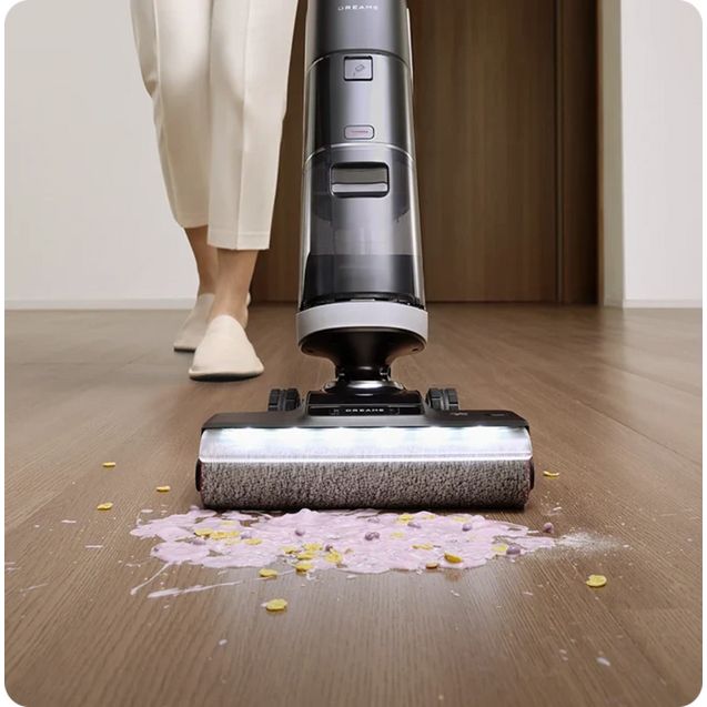 Dreame H14 Pro wet and dry vacuum cleaner in black and silver actively cleaning scattered petals and liquid from a wooden floor, with a person wearing white clothing and slippers standing behind the machine