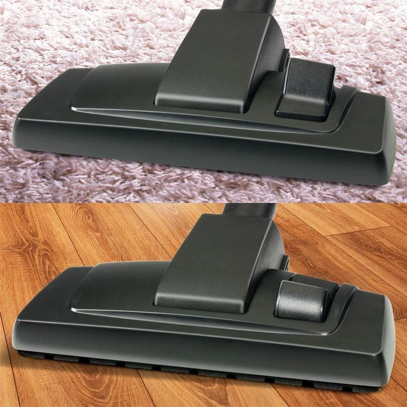 Vacuum cleaner floor brush head attachment shown on pink carpet and wooden flooring surfaces, demonstrating dark gray motorized cleaning head with rotating brush roll and adjustment controls