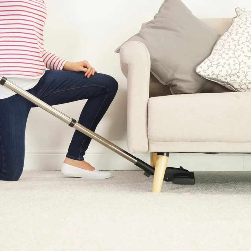 Person using a Henry vacuum cleaner with a long handle to clean beige carpet under a cream upholstered sofa with gray and patterned pillows, wearing pink striped top and dark jeans