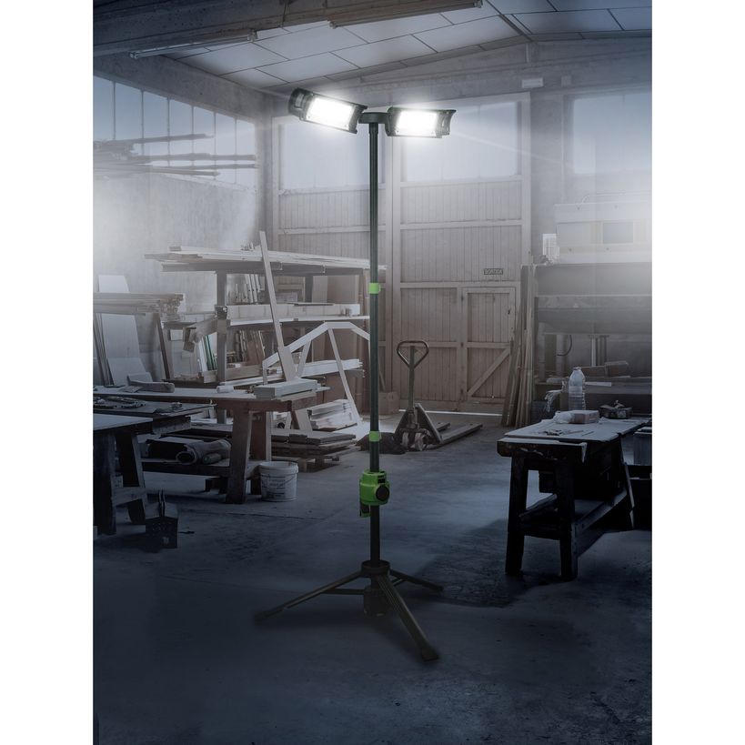 Lighthouse rechargeable site light with dual LED heads mounted on adjustable green and black tripod stand illuminating industrial workshop garage setting with wooden materials and workbenches