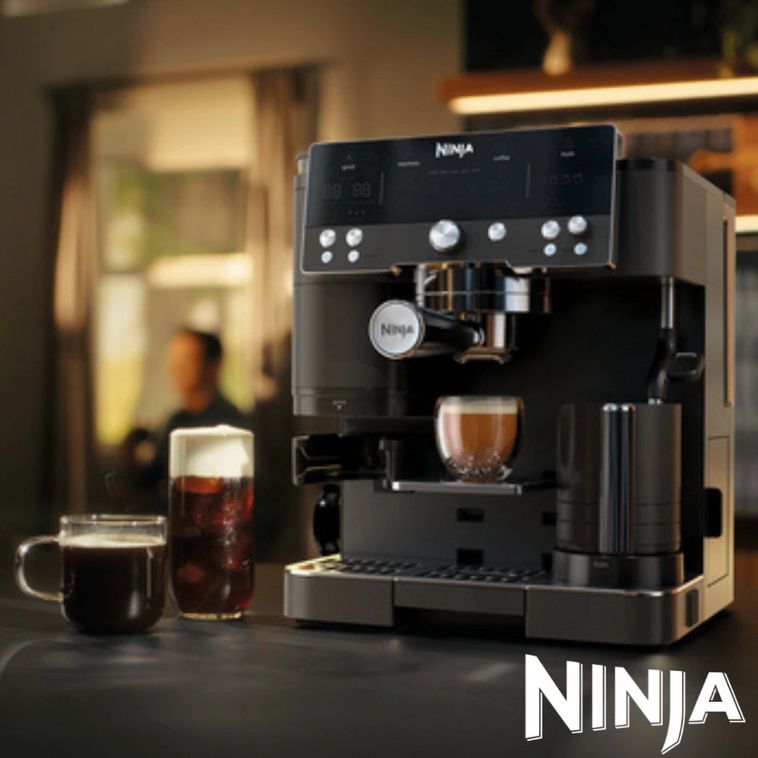 Ninja Luxe Café Premier Series black espresso machine with stainless steel accents dispensing coffee into a cup, displayed with a black mug and cold beverage glass on a dark counter in a modern kitchen setting