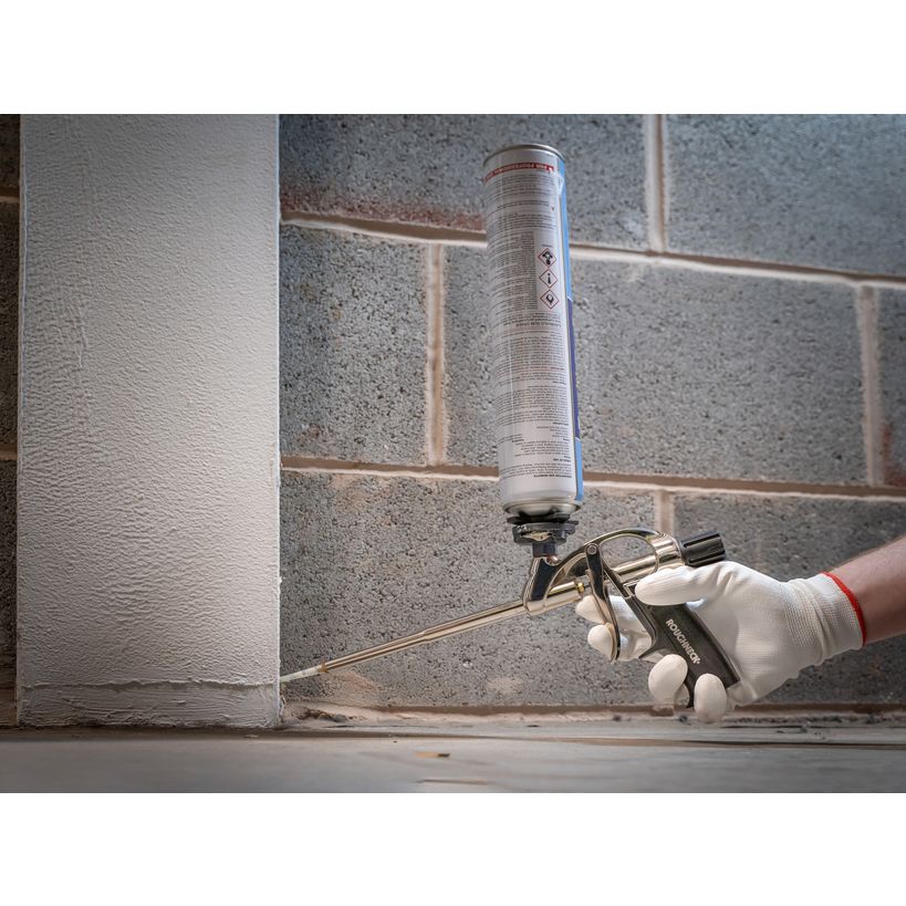 Worker in white gloves holding Roughneck expanding foam gun with cylindrical cartridge against concrete block wall during application