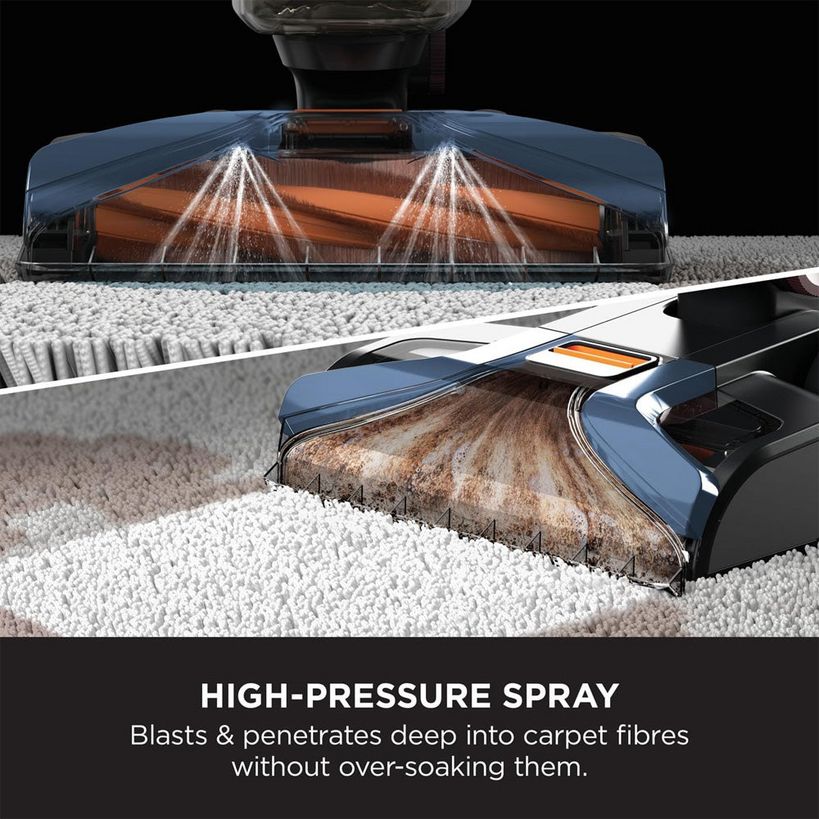 Shark CarpetXpert deep carpet cleaner with navy blue body demonstrating high-pressure spray technology cleaning light gray carpet fibers, showing dual spray nozzles and cleaning brush head in action