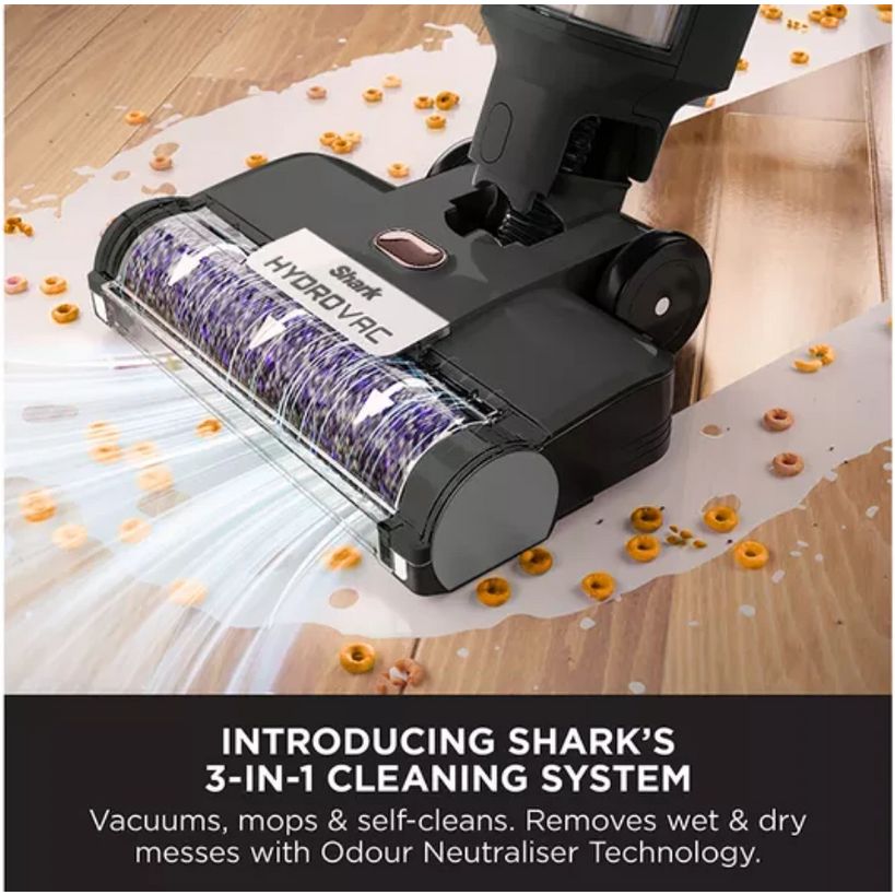 Shark HydroVac cordless vacuum cleaner demonstrating 3-in-1 cleaning system vacuuming cereal pieces and crumbs from wooden floor with white surface, black motorized cleaning head with purple brush roll visible, product features wet and dry cleaning with odour neutraliser technology