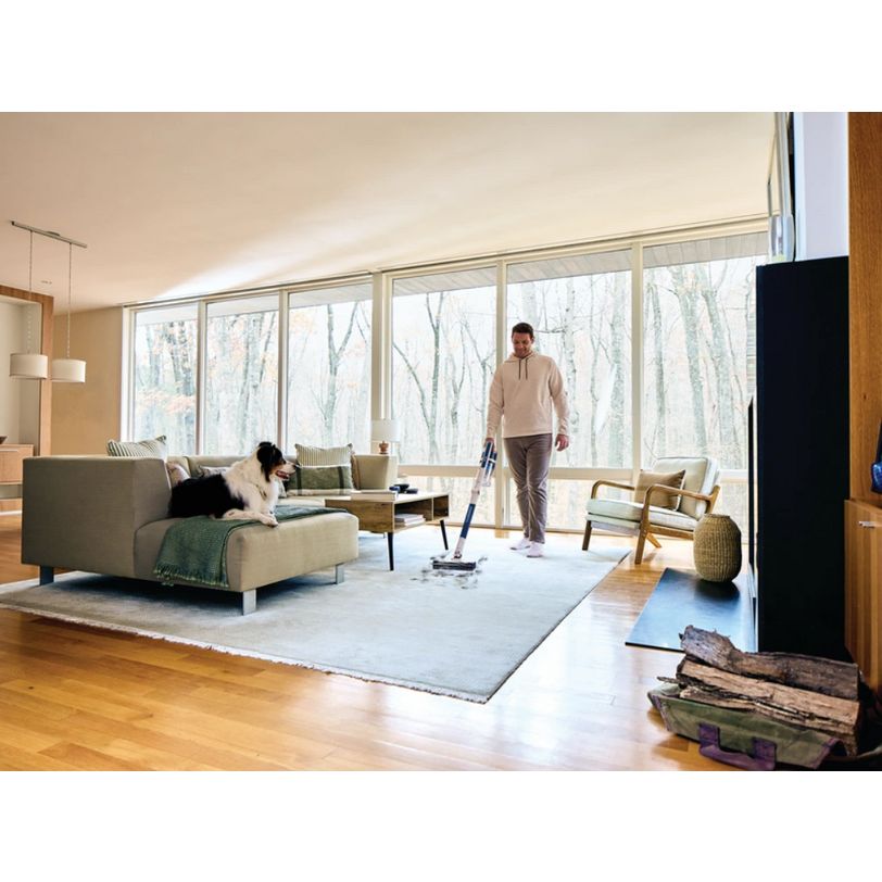 Man using Shark PowerPro cordless stick vacuum in modern living room with beige sofa, wooden flooring, large windows overlooking forest, and woman relaxing on couch