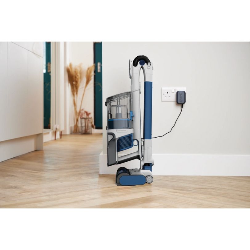 Shark PowerPro cordless stick vacuum in white and blue with anti hair wrap technology, displayed on wooden flooring in a modern minimalist home interior, showing the charging dock on the white wall behind it