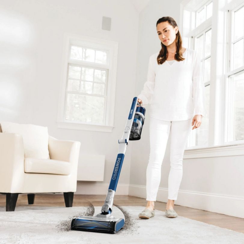 Woman in white clothing demonstrating the Shark PowerPro Cordless Stick Vacuum with blue and black design on a light grey rug in a modern living room with cream armchair and white walls