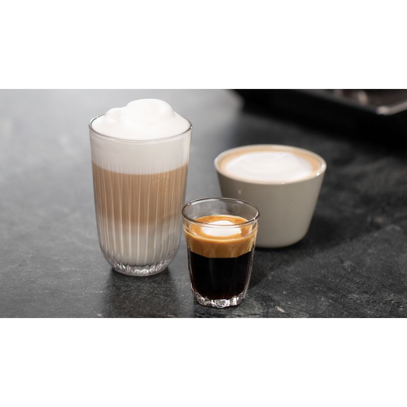 Three espresso-based coffee drinks displayed on a dark gray surface: a tall glass with cappuccino topped with thick white foam, a small glass of dark espresso with crema, and a white ceramic cup with flat white or latte